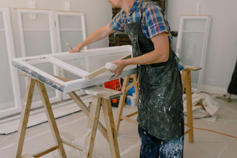 Unrecognizable male master wearing dirty apron standing near window with white frames on wooden workbench while working in professional studio